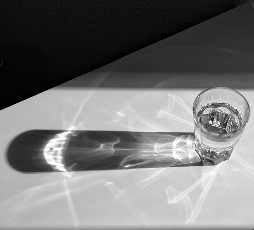 Das Bild zeigt ein Glas Wasser auf einem weißen Tisch. Das Glas ist halb gefüllt und steht aufrecht. Die Sonne scheint auf das Glas und erzeugt ein komplexes Lichtspiel auf dem Tisch, das die Form des Glases und die Struktur des Wassers widerspiegelt. Die Schatten und Lichtreflexe erzeugen ein kunstvolles Muster, das die Struktur des Glases und die Transparenz des Wassers hervorhebt. Der Hintergrund ist dunkel, was den Fokus auf das Glas und das Lichtspiel lenkt. Die gesamte Szene ist in Schwarz-Weiss gehalten, was die Kontraste und Texturen verstärkt.