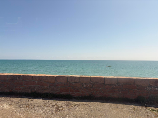 A stone wall overlooks a vast ocean under a clear blue sky with a small boat in the distance.