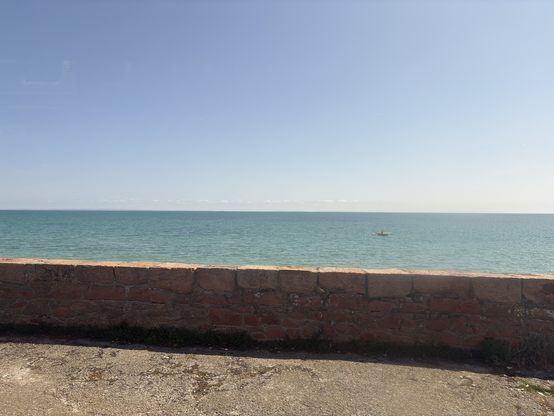 A stone wall overlooks a vast ocean under a clear blue sky with a small boat in the distance.