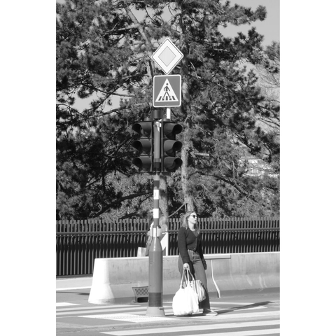Black and white street photography taken in Luxembourg city on the Boulevard Roosevelt. A lady with sunglasses is waiting at the traffic light. She wears big bags and seems exceeded to wait in the sun, maybe for a long time. Road signs on top of the traffic light make it look like a totem to which the lady would be tied.