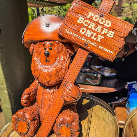 A wooden bear figure wearing a cowboy hat holds a sign that reads "FOOD SCRAPS ONLY" and "This bin is for feedin' critters!" The bear is sitting on a table, with some people and food trays visible in the background