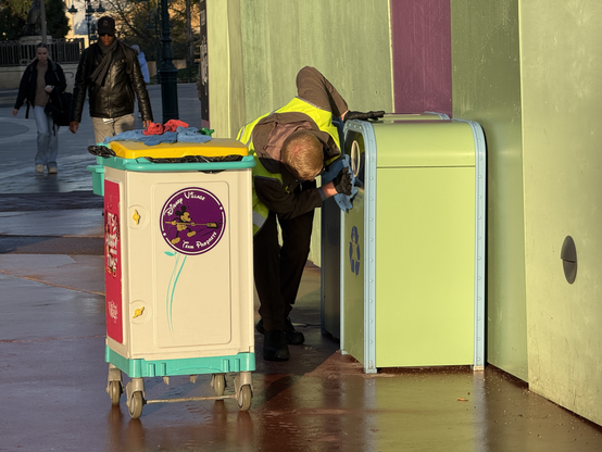 A Disney Village custodial cast member in a safety vest is cleaning a green recycling can, while a colorful trash can on wheels is nearby. The scene features pedestrians walking in the background, reflecting a lively environment. 

