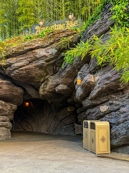 A rocky entrance to Adventure Isle, featuring a sign above, surrounded by lush greenery and bamboo. Two decorative trash cans are positioned on the right side. Soft lighting is provided by hanging lanterns along the entrance.