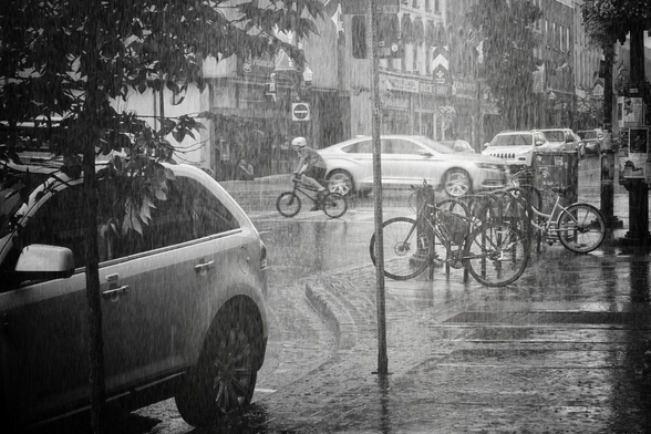 A cyclist crosses through an intersection during a heavy rain storm. Cars are seen on the road and parked at the curb, trees are seen on the sidewalk and numerous bikes are locked up. This is a black and white image.
