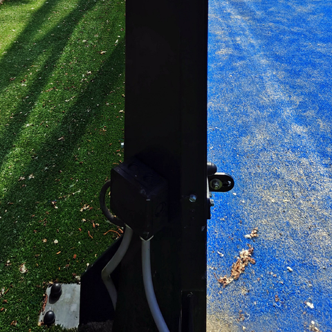 A close-up of a black metal post with some electrical wiring near the bottom that stands between two surfaces: green artificial turf on the left and blue synthetic ground on the right.

