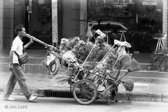 A Simple And Mundane Black And White Street Photo In Landscape Format. In This Street Scene An Oriental Man Is Walking From Left To Right On The Road And Pushing A Small Wheeled Cart Stacked With A Few Household Brooms And A Great Many Very, Very Fluffy Feather Dusters, A Few Of These Dusters Fixed To Two Metre Or Longer Bamboo Poles. Our Feather Duster Executive Has Medium Length Dark Curly Hair, Is Wearing A Short Sleeved White Shirt, A Man Bag Over His Shoulder And Resting On His Chest, Jeans (Torn At The Right Knee And Trainers. Behind Is A Pavement With An Empty Bar (Because It Has A 'Closed' Sign On The Entrance) Beyond. 
The Location: Bangkok, Thailand. 
