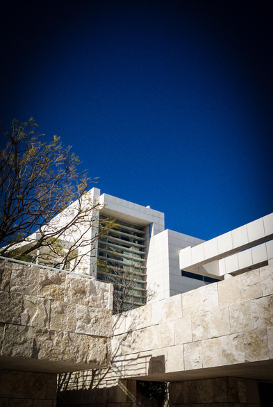 The image features a modern architectural structure set against a clear blue sky. The building is predominantly white, with a combination of smooth and textured surfaces. The facade includes large glass windows, allowing for natural light to enter. The structure has a geometric design with clean lines and angular forms, including a prominent overhanging section. In the foreground, there is a stone wall with a rough texture, contrasting with the smoothness of the building. A tree with sparse branches is visible on the left side, adding a natural element to the scene. The overall composition highlights the interplay between the man-made structure and the natural environment.

Provided by @altbot, generated privately and locally using Ovis2-8B