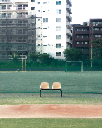 Two cream-colored chairs on a metal frame positioned on a track, facing a fenced soccer field. The field has a goalpost and is partially enclosed with netting. In the background, there are high-rise buildings, some with scaffolding, surrounded by greenery.