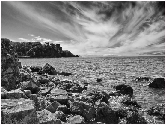 A black and white landscape featuring rocky shoreline, with water in the foreground and a distant cliffside castle surrounded by trees under a cloudy sky.