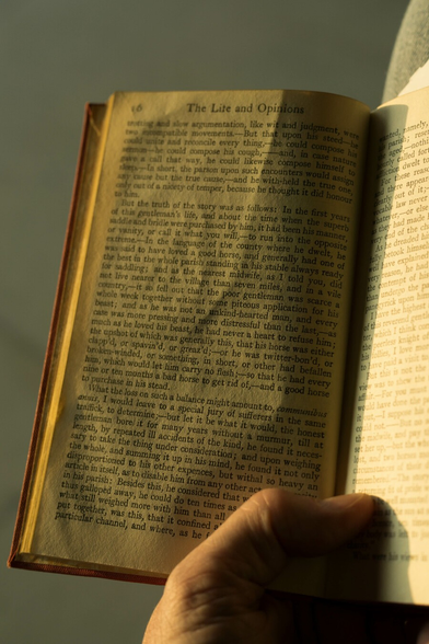 Gently sunlit photo of the left-hand side of an old open book, the red cover visible around the yellowing, slightly rumpled, pages. It's held open by a left hand, only the thumb of which is in shot.