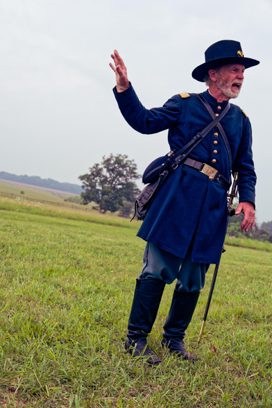 Man in blue blue coat and hat with white bear is talking, wearing a sword and boots standing on a lawn with a slanted horizon behind him.