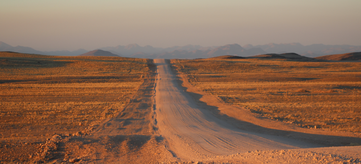 in the center of the photo, a dirt road stretches into the distance and to a hill, beyond which it disappears. the road cuts through a flat desert, and mountains can be seen in the hazy distance as he sun sets from the opposite direction, creating a pastel sky