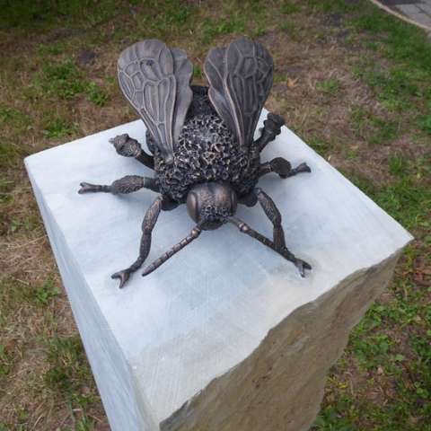 Skulptur einer Stubenfliege aus bräunlichem Metall auf einer hellen Steinplatte. Die Figur ist übergroß und zeigt filigrane Details.