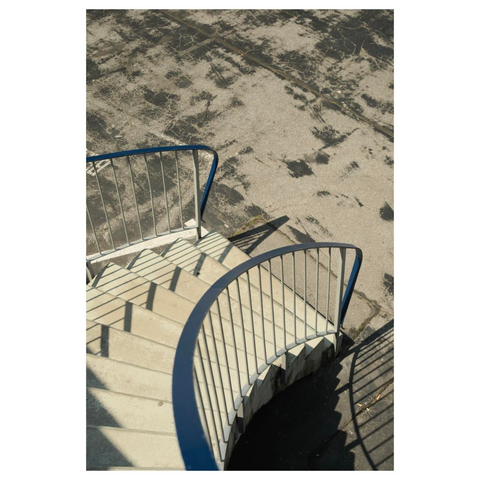 Block von oben herab auf eine geschwungene 60er Jahre Betontreppe. Weiße, vertikale Streben halten einen blauen Handlauf, der scharfe Schatten auf den fleckig geteerten Parkplatz wirft, auf dem die Treppe endet. 