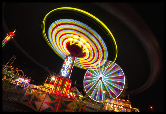 An 11 second exposure of carnival rides, taken at night. Two brightly lit, extremely colorful rides fill the center of the photo, set against black skies. Furthest away from the camera is a large ferris wheel, with multi-colored spokes. And closest to the camera is a ride called Yoyo. Yoyo is a spinning wheel mounted on top of a center column. Baskets of people hung from Yoyo's wheel are flung through the air in a circular orbit when Yoyo starts spinning. And as people spin, blood rushes from their heads to their feet. Solids are separated from liquid. Heavy metals, in particular, migrate to the toes.

And people pay to do this.