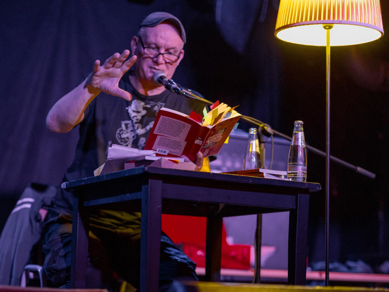 A man wearing glasses and a cap is reading from a book while gesturing with one hand. He is seated at a table with a microphone, a lamp, and bottles of water. The background is dark, indicating an intimate setting.