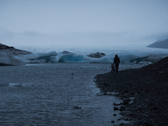 Mother and child walking hand-in-hand on the rock beach in the distance. Behind them there is large blue ice blocks from glacier in Iceland. The weather is chilly and misty.
