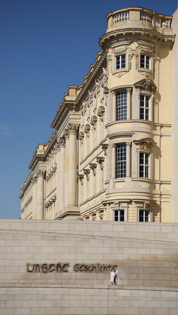The image shows a historic building with ornate architectural details in the background. In the foreground, someone is cleaning or painting over spray-painted graffiti that reads "UNSERE Geschichte" - „our history“ on a stone wall. The sky is clear and blue.