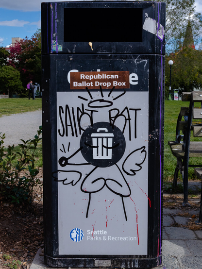 Image of a black metal trash receptacle labeled with "Seattle Parks & Recreation" and its logo. The garbage can has some graffiti on it, with a solid black circle with an icon of a trash can being used as the head of a drawing of a rat with angel wings. Beneath it's glowing halo, the words "SAINT RAT" seem to give the cartoon a name. On top of the word "GARBAGE" that would usually don the bin, a brown sticker with the text "Republican Ballot Drop Box" has been placed.