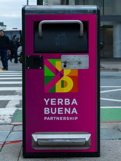 Image of a purple & pink trash can. The bin has a panel with the words "Yerba Buena Partnership" and a green, yellow, and orange logo.