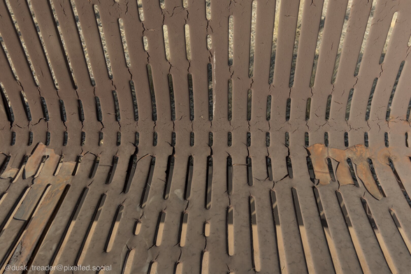 An overhead view of a weathered metal bench with visible cracks and wear, featuring elongated slats and rust patches. The background is blurred and shows a gravel surface.