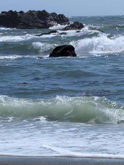 Some waves forming crashing rolling on rocks, a blueness above and a little foamy beach below.