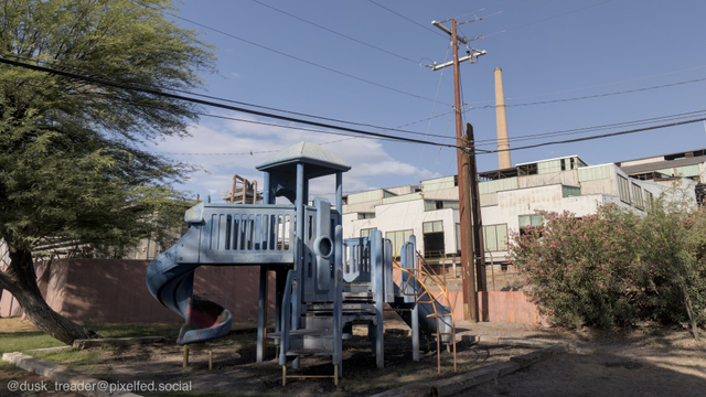 A blue playground structure with a slide and climbing features is situated in an open area, surrounded by power lines and industrial buildings in the background. A tree and some shrubs add greenery to the scene.