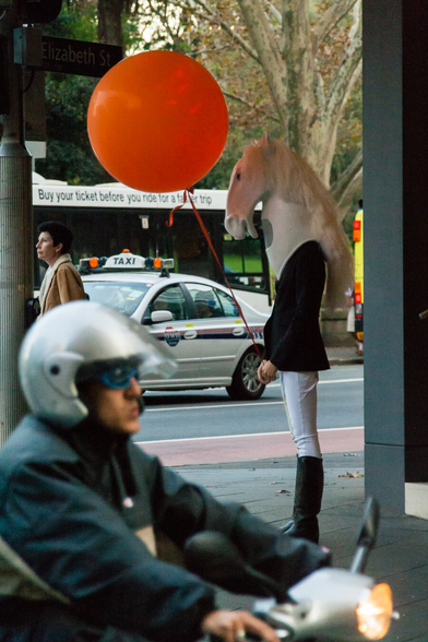A person wearing a horse head costume and holding a red balloon is standing on the corner of a street in the Sydney city centre, apparently advertising something for  a shop. At the same time, a motorcycle rider with helmet is driving past on the lower left. In the background there's a bus at a bus stop.
