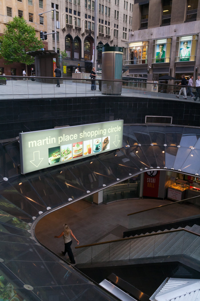 The lower two thirds of this photo shows steps leading down to an underground shopping area in a train station in the centre of Sydney. A woman is walking down below, and the sign above says Martin Place Shopping Circle. In the upper third of the image is the street level, with commercial buildings in the background.