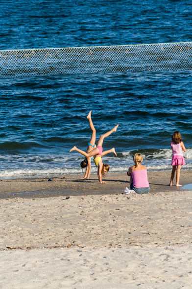 Two young girls are standing on their hands on the sand in front of the waves on a beach. They are almost body to body with their heads facing outwards. To the right of them, a woman is sitting and a young girl is standing. They're both facing the water. In the foreground of this photo, there just sand, andabove that, only water and part of a shark net. This could be taken anywhere, as there is nothing else to identify the place.