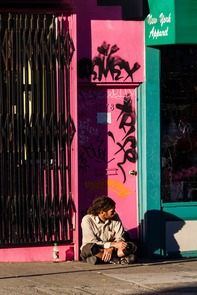 A young man is sitting in the doorway of a building in the Haight-Asbury neighbourhood in San Francisco. He is looking to the right, with his face nearly in profile, and he has dreadlocks tied back neatly in a short ponytail. There's an empty plastic drink bottle to his left.  The door has graffiti on it. At the top right, in the green area, other words New York Apparel.