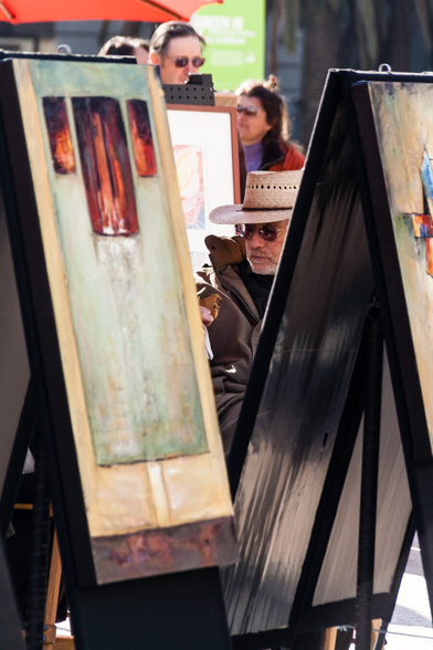 The face of a middle-aged looking painter wearing sunglasses and a straw hat peeks out between paintings which appear to be on easels in San Francisco's Union Square. He appears to be concentrated and working on something. The heads of a couple of other people can be seen in the background.