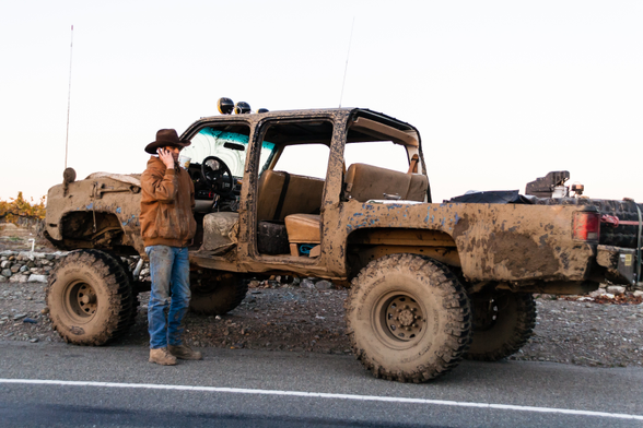 A man in a cowboy hat, jacket and weathered looking jeans stands on the side of a road in front of doorless, paint scarred, battered looking Jeep type pickup truck with large wheels and raised suspension. He has his mobile phone to his ear. The background is a completely colourless sky.