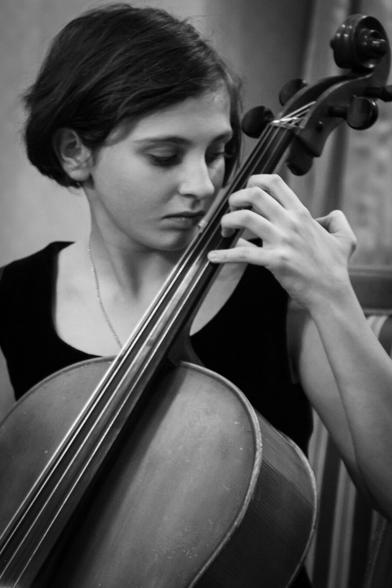 Black and white photo of a young woman with short dark hair playing the cello. Photographed from the front. You can only see her face and her hand on the upper part of the strings and the upper part of the cello. She is looking concentrated at her the strings. The hand is slender and bony.