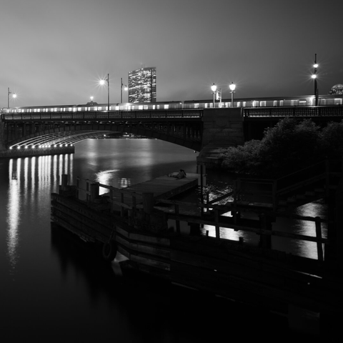 Black & white photo of the Cambridge end of the Longfellow bridge. There's a stopped train at the top, and below is a wooden dock with a couple sitting.