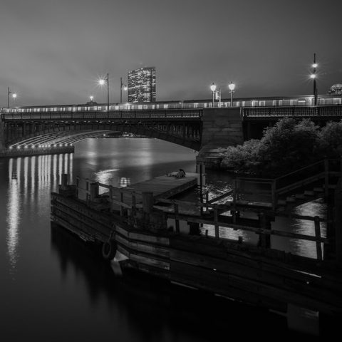 Black & white photo of the Cambridge end of the Longfellow bridge. There's a stopped train at the top, and below is a wooden dock with a couple sitting.