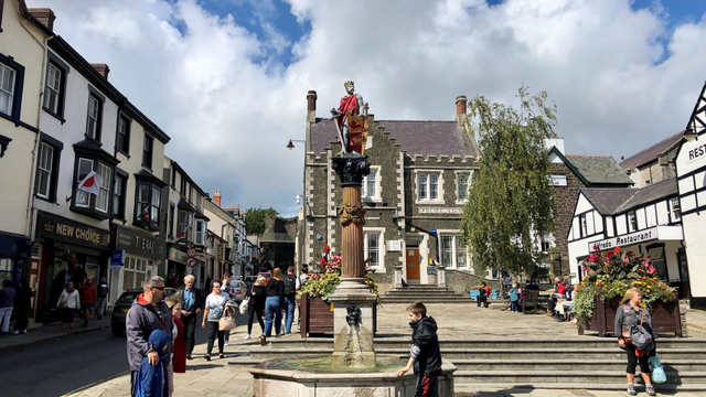 The photo depicts a lively street scene in a charming town square, likely in Wales, given the flags and architectural style. At the center is a tall statue of a crowned figure holding a shield with a red lion emblem, standing atop a decorative column. At the base of the column is a fountain with water spouting from a sculpted lion's head. Surrounding the square are traditional buildings with white facades, black timber framing, and stone structures. Shops like "NEW CHOICE" and "T E R A J" are visible on the left, alongside a restaurant named "Alfredo Restaurant" on the right. The area is bustling with people walking, shopping, and relaxing, with flower arrangements adding vibrant color. The sky is partly cloudy, casting a bright, cheerful light over the scene.