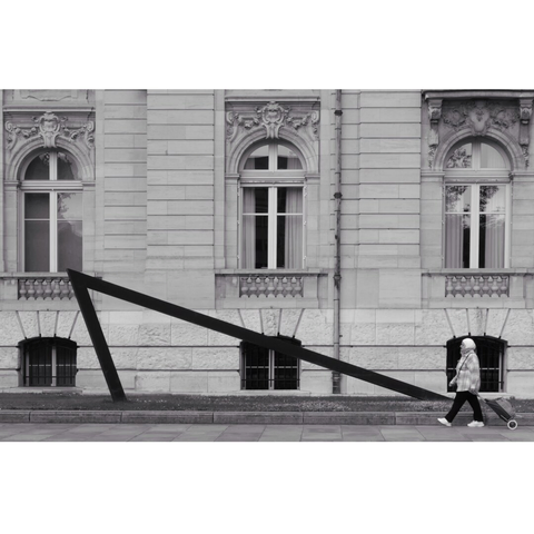 Street photography taken in Luxembourg city. The background is the Arbed building. A lady drags a trolley as she walks, on the right corner of the image. She is at the lower right of the triangle line made of steel and placed there a few years ago to remind the past of what houses now a bank.