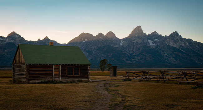 A log cabin with a green roof next to a wooded fence with craggy mountains in the background.