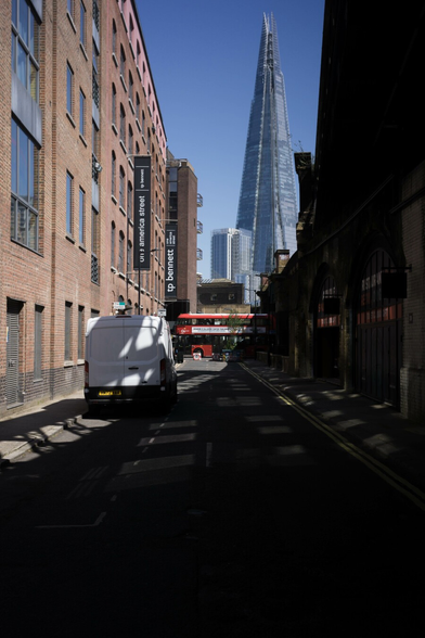 Photo looking down a street in deep shade on a clear bright day, the darkness falling across from a raised railway on the right. On the left a white van touched by shadow is parked in front of what looks like an old brick warehouse extended and converted into offices. At the end of the street a red double-decker bus passes by. In the distance are two buildings, glass and steel, faded by distance and shaded blue by the sky. The shorter, probably around twenty stories, is boxy and dwarfed by the spike of The Shard, looking even more than normal like an intruder in a science fiction film.