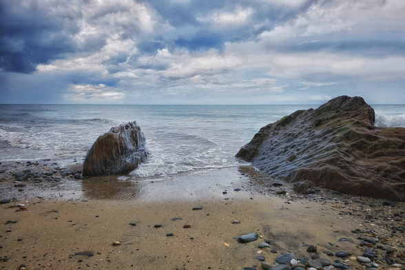 A dramatic coastal scene featuring two large, dark rocks on a pebble and sand beach. The ocean stretches out to the horizon under a heavily clouded sky. Waves gently lap against the shore and around the base of the rocks.