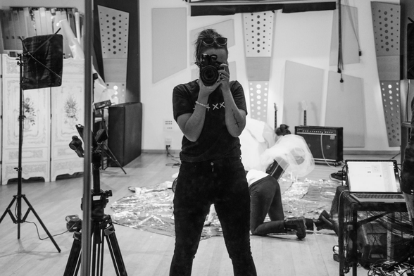 A black and white self portrait shot through a mirror in a rehearsal space. In the background a band can be seen setting up the room for a makeshift video shoot