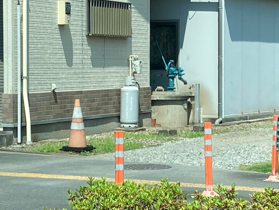 An image showing a gray building facade with a light-colored section, a blue water pump on a concrete platform, a propane tank, a traffic cone, orange traffic markers, and some greenery in the foreground.