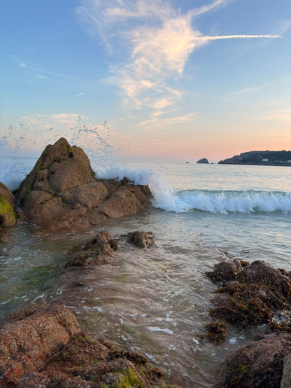 Waves crash against rugged rocks on a peaceful shoreline under a sky with wispy clouds at sunset.