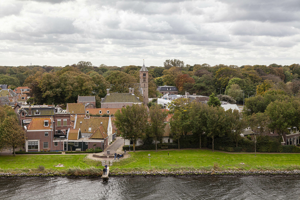 We're on the balcony of a medium-sized cruise ship, about halfway up its height, and the ship is currently slowly heading along a wide canal cutting through European countryside. The view of the land is largely trees filling the background, still leafy but with autumnal colours, and nestled in amongst them nearer the water is a small village or town of houses and a church tower. Red brick dominates the building material used and the tiles are dull browns, oranges, or pale reds for the most part. A very small stone jetty emerges from the bank leading off a small pedestrianised area between two rows of houses and there are a few people gathered there, one with a tripod set up to capture the sight of our ship sailing past probably quite a sight in this local environment. It's a cool, cloudy day.