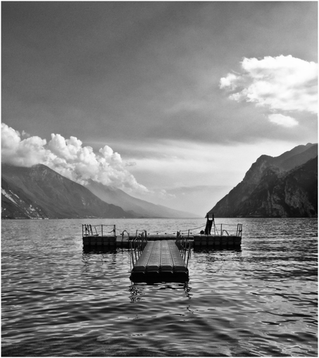 A black and white image of a pontoon on a calm body of water, surrounded by mountains and a cloudy sky.