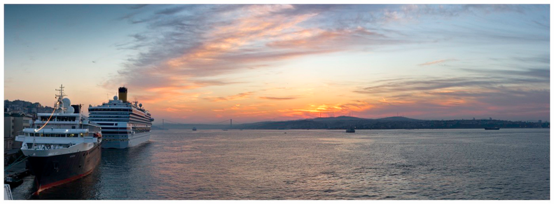A panoramic view of a harbor at sunset, featuring two cruise ships docked beside each other. The sky displays vibrant colors with clouds, while the water reflects the sunset hues. A bridge is visible in the background along with hills and city buildings.