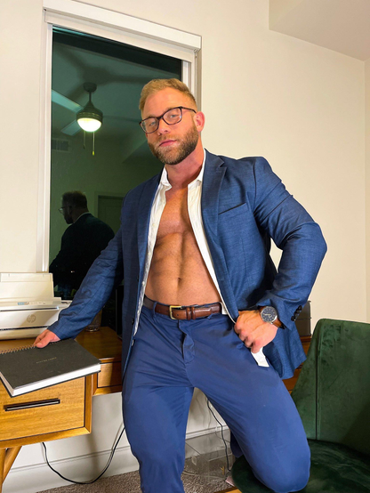 A muscular white man wearing a blue suit and glasses is looking at the camera with his unbuttoned white shirt, showing his muscled torso