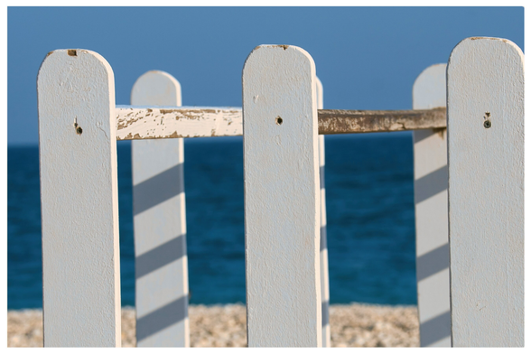 Nahaufnahme, fast formatfüllend eine weiss lackierte Holzkonstruktion mit leichten  Abplatzungen, dahinter unscharf Kieselstrand, dunkelblaues Meer und Himmelsblau.
