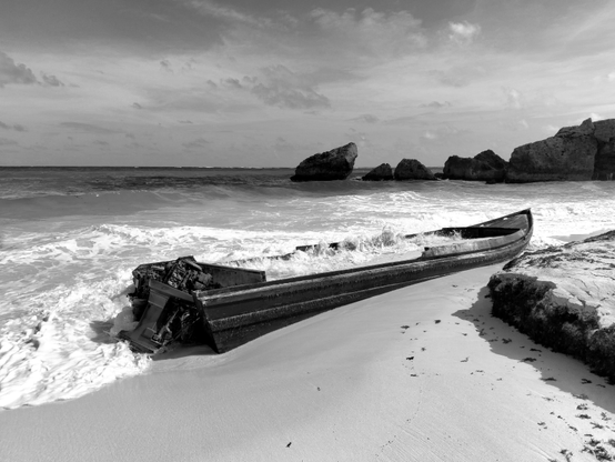 The image depicts a black and white scene of a beach with a large, weathered boat partially submerged in the shallow water. The boat, which appears to be a traditional wooden vessel, is lying on its side on the sandy shore, with waves gently lapping at its hull. The boat's structure is visibly deteriorated, with parts of the wood and metal framework exposed, suggesting it has been abandoned for some time. The beach is sandy, with a few scattered footprints leading towards the boat, indicating recent human presence. In the background, the ocean extends to the horizon, with several large rocks protruding from the water, adding to the rugged coastal landscape. The sky is partly cloudy, with soft, diffused light illuminating the scene, enhancing the textures of the sand, water, and boat. The overall composition emphasizes the contrast between the natural elements and the man-made object, highlighting the boat's state of disrepair against the backdrop of the sea and sky.

Provided by @altbot, generated privately and locally using Ovis2-8B

🌱 Energy used: 0.217 Wh